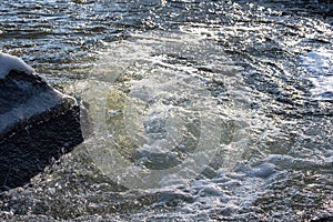 The rapid flow of the river breaks on a large stone covered with ice. The water froths and sparkles in the spring sun photo