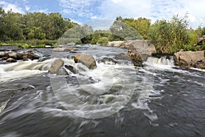 The rapid flow of the river, rocky coasts, rapids, bright green vegetation and a cloudy blue sky in summer - a frontal view