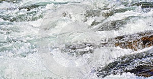 Rapid spring river flowing over rocks on sunny day, forming white water waves, closeup detail - abstract nature background