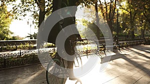 Rare view of a young man in black casual clothes, headphones riding a bike by the paved city park with guitar in black