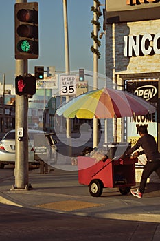 Raspado Man pushes red cart with rainbow umbrella down the street