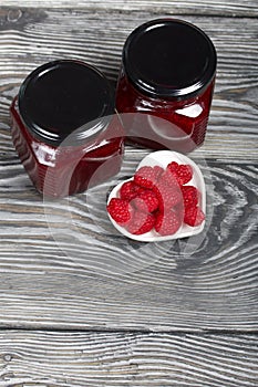 Raspberry jam in glass jars. Nearby are raspberries in a saucer. On wooden boards with a beautiful texture