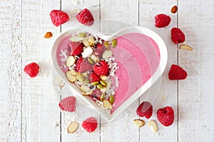 Raspberry smoothie in a heart bowl with superfoods, above on white wood