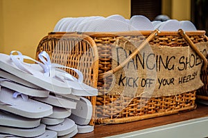 Rattan basket with white flip-flops of different sizes for guests, with a writing DANCING SHOES. KICK OFF YOUR HEELS!