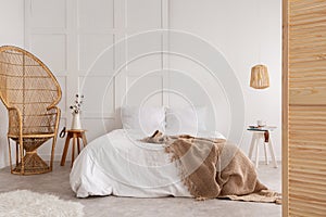 Rattan chair and wooden table next to bed with brown blanket in white bedroom interior. Real photo