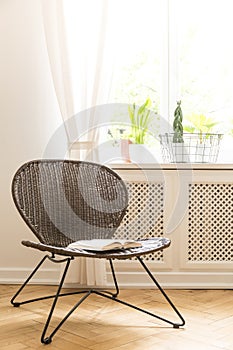 A rattan and metal chair with an open book on a seat standing on a wood flooring against a white wall and a sunny window in a livi