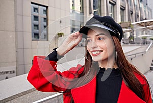 Ravishing luscious delightsome girl in red overcoat holds her kepi by hand, looks into the distance and smiles