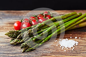 Raw asparagus stems with a brunch of cherry tomatoes and a pinch of coarse sea salt on a rustic wooden table with black background