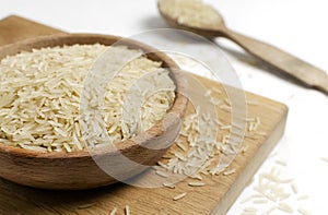 Raw basmati rice in a wooden bowl with a spoon on a cutting board.