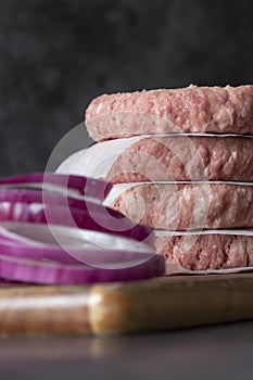 Raw beef burgers stacked on a wood cutting board, with greaseproof paper. With red onion in the foreground