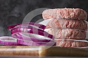 Raw beef burgers stacked on a wood cutting board, with greaseproof paper. With red onion in the foreground