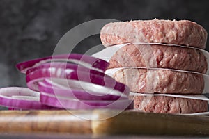 Raw beef burgers stacked on a wood cutting board, with greaseproof paper. With red onion in the foreground photo
