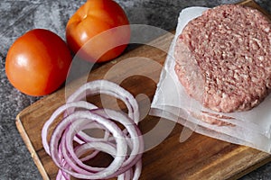 Raw beef burgers stacked on a wood cutting board, separated with paper. With red onion and tomatoes