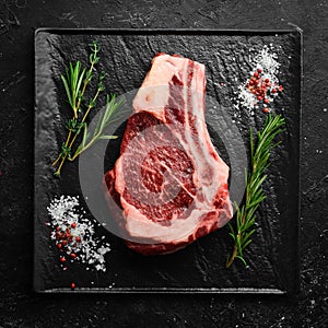 Raw beef ribeye steak on the bone with thyme, rosemary and spices.  Flat lay top view on black stone cutting table