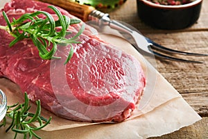 Raw beef steak. Fresh beef rib eye steak with fork rosemary, salt and pepper on piece of parchment paper on old wooden background.