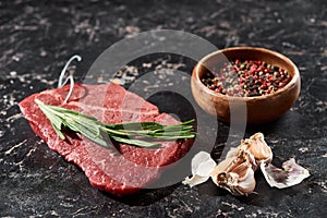 Raw beef steak with rosemary twig near small bowl with peppercorns mix and garlic on black marble surface.