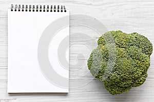 Raw broccoli and blank notepad on white wooden table, overhead view.