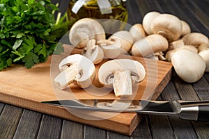 Raw champignon cut in half and and kitchen knife on a cutting board, whole button mushrooms and bunch of parsley over dark