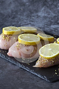 Raw chicken breast fillets, covered with rosemary and sliced lemon, on a slate cutting board.