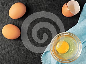 Raw chicken egg in a glass cup, dark stone background. Top view of a bright yolk, next to an egg shell, two whole eggs. The