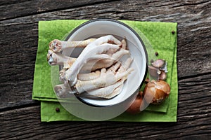 Raw chicken legs lie in a bowl on a wooden table. Near spices and onions with garlic. Green kitchen napkin under the plate. Useful