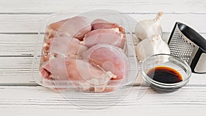 Raw chicken thighs in a plastic container, soy sauce, and fresh garlic and garlic press close-up on a white wooden board