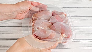 Raw chicken thighs in plastic container in woman's hands close-up on white wooden background