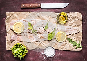 Raw cod with lemon and herbs, salt and butter and a knife on paper on wooden rustic background top view close up