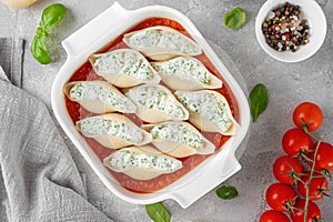 Raw conchiglioni pasta stuffed ricotta cheese and spinach with tomato sauce in a white baking dish on a gray concrete background