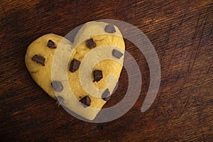 Raw cookie dough with chocolate chips in the shape of a heart on a wooden background.
