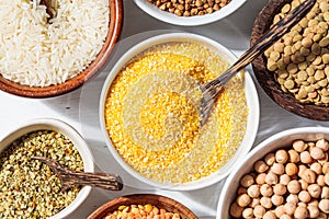 Raw corn grits in white bowl. Various dry cereals and legumes in white and wooden bowls, white wooden background. Vegan protein