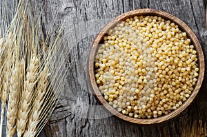 Raw couscous in bowl on wooden background with wheats