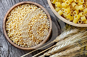 Raw couscous in bowl on wooden background with wheats