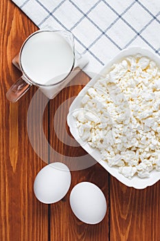 Raw curd cheese in a white bowl. Ingredients on a brown wooden table for cooking. Eggs, cottage cheese and milk on a board, food