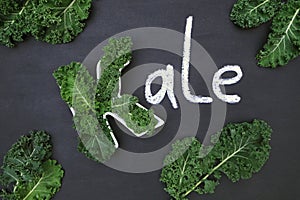 Raw curly-leaf kale cabbage inside letter K shaped plate, chalk inscription KALE and fresh green leafy vegetables on blackboard