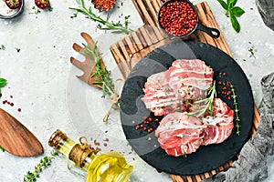 Raw cutlets in bacon served on a black plate with rosemary and thyme. On a gray background.