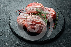 Raw cutlets wrapped in bacon served on a black plate with rosemary and thyme. On a stone background.