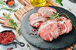 Raw cutlets wrapped in bacon served on a black plate with rosemary and thyme. On a stone background.