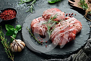 Raw cutlets wrapped in bacon served on a black plate with rosemary and thyme. On a stone background.