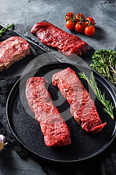 Raw denver, top blade, tri tip steak on a black plate and stone slate with seasonings, herbs grey concrete background. Side view