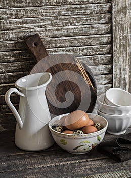 Raw eggs in a bowl, enamelled jug, chopping board and ceramic bowl on a rustic dark wooden background.