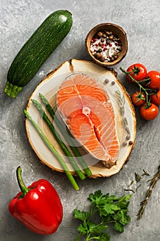 Raw fish steak salmon with fresh vegetables, asparagus, zucchini, sweet peppers, tomatoes, herbs and spices on a gray background.