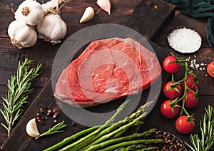 Raw fresh beef braising steak on chopping board with garlic, asparagus and tomatoes with salt and pepper on wooden background