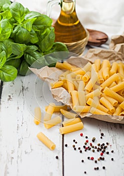 Raw  fresh homemade penne pasta in brown paper with basil and oil on white wooden background