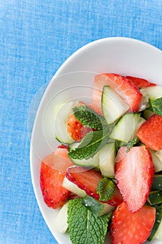 Raw fruit detox salad with cucumbers strawberries apples fresh mint in crystal bowl on blue table cloth background. Vegan