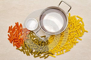 Raw fuzilli with water in pot and salt. Multicolored pasta scattered over on stone background. Preparing for children, top view,