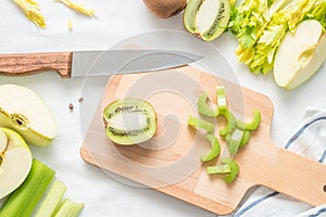 Raw green ingredients for smoothie, whole and cut apples, chopped celery stalk on a wooden board on white cloth background