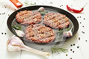 Raw hamburgers - cutlets from organic beef meat with garlic, chilli and rosemary in a frying pan on a white background