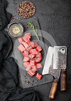 Raw lean diced casserole beef pork steak on chopping board with vintage meat hatchets on stone background. Salt and pepper with