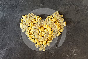 Raw macaroni - pasta Conchiglie. In the middle of the empty space in the shape of a heart on a dark concrete background.